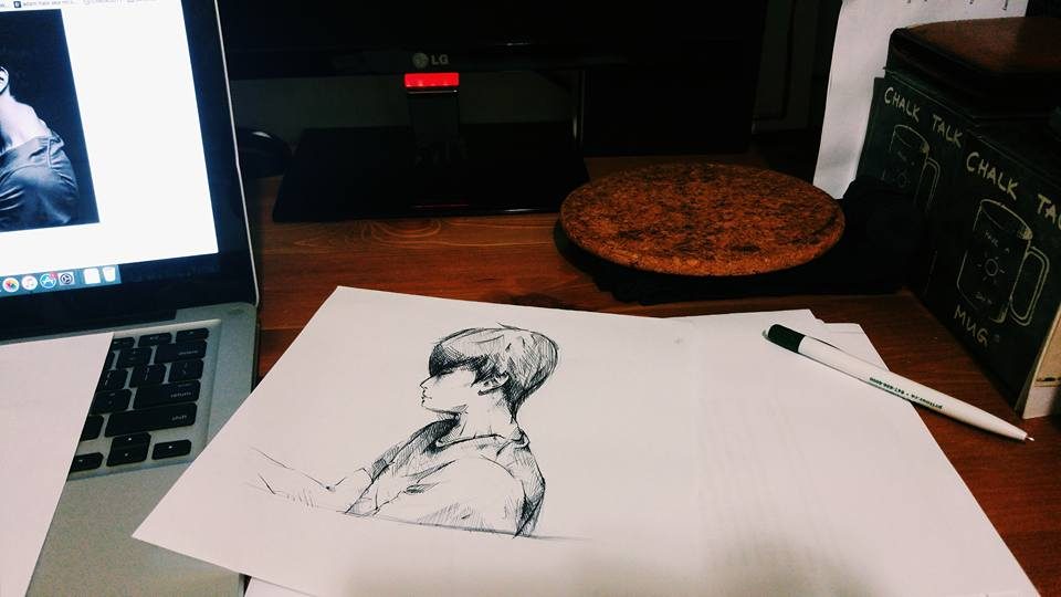 A pencil portrait sketch on a desk beside a laptop